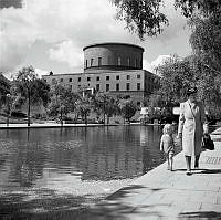 Observatorielunden. Kvinna och barn går hand i hand längst med plaskdamm. Stadsbiblioteket i bakgrunden.