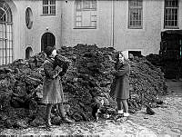 Två flickor med skridskor på fötterna leker med en stor hög av torv. Torven eldades som reservbränsle.