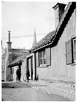 Två barn står vid porten till Iversons Gränd 5, nu Iversonsgatan. Pojken håller i ett tunnband. I fonden västerut ses tornet på Johannes kyrka.