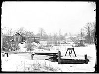 Byggnader och brygga i området kring sjön Trekanten. I fonden skorstenar vid Palmkrantzka fabriken i Lövholmen