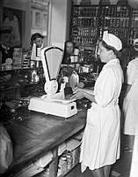Brödransonering under andra världskriget. Kvinna i vit rock i mataffär väger bröd på en våg åt kunder.