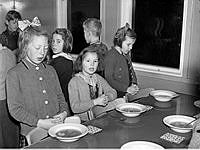 Barn vid matbord i skolbespisning står upp med knäppta händer och läser bordsbön.