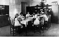 Interiör från Föreningen Engelbrekts barnavärn och husmodersskola med barn och personal som sitter runt ett bord och läser.