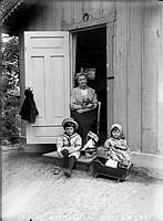 Kvinna  och två barn framför ingången till en stuga i skärgårdsmiljö.
