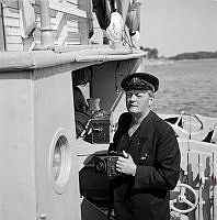 Reportage från Sandhamn under sandhamnsregattan 1941. Med en fläkt från sol, sommar och Sandhamn. En man ombord på en båt med kamera och SSB (Segel Sällskapet Brunnsviken) märke i kepsen.