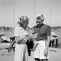 Reportage från Sandhamn under sandhamnsregattan 1941. Den nordiska seglarfesten.
Med en fläkt från sol, sommar och Sandhamn. Två kvinnor tittar i en broschyr i hamnen.
