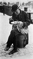 En försäljare av ståltrådsarbeten sitter på en stenpollare med sina varor uppträdda på armen och äter mat från en tallrik.