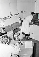 Interiör från kök på Rålambsvägen i Marieberg. En kvinna från Hemmens forskningsinstitut gör tidsstudier, med klocka och anteckningar, på en kvinna som arbetar vid köksbänken i köket.