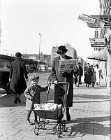 Tidningsläsning på stan. En kvinna promenerar på Götgatan med barnvagn samtidigt som hon läser tidningen. I fonden kvarteren Gamen och Nattugglan.