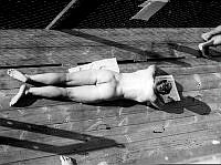 Läsande naken man ligger och solbadar på en brygga.