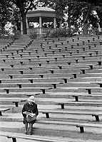 En kvinna sitter på en bänk på Friluftsteatern i Vitabergsparken. I bakgrunden skymtar musikpaviljongen.