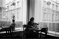 Norra Smedjegatan 16. Interiör från Linds kaffestuga. En kvinna sitter vid ett cafébord och läser en tidning. Genom gardinerna skymtar del av kvarteret Torviggen.