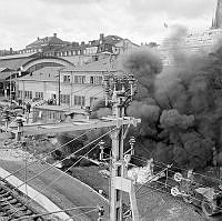 Explosion och brand i en asfaltsbehållare på centralbangården vid Centralen. Svarta rökmoln. Män på tak och i fönster.