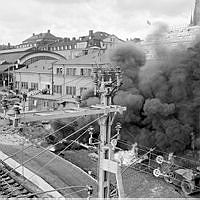 Explosion och brand i en asfaltsbehållare på centralbangården vid Centralen. Svarta rökmoln. Män på tak och i fönster.