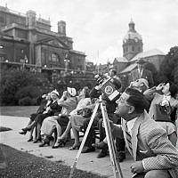 Reportage under solförmörkelse. Folk tittar på solförmörkelsen. En man filmar framför Operakällaren. 