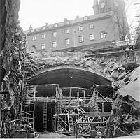 Vy från Klara Vattugränd. Byggnadsställningar vid ett sprängt hål för tunnelbanan. Huset ligger på Klara Västra Kyrkogata, kvarteret Klockstället. I bakgrunden Klara kyrka.