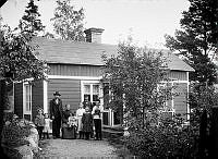 Gruppbild av två män och och en kvinna, sex barn och en katt, framför ett hus.