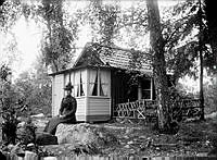 En kvinna sitter på berget framför ett litet hus. 