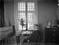 Konstnären och författaren Albert Engström sittande framför ett staffli. På bordet ligger en bunt med Söndags-Nisse. På byrån en Hasselblad Express-Kamera.