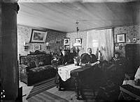 Sittande runt ett bord syns tullvaktmästare Hjalmar Robert Öhman, Anna Maria Carlsson  och pigan Agda Sofia. Kudden i soffan har kungliga tullverkets monogram.