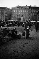 Torghandel på Kornhamnstorg. En kvinna med paket kommer gående. Träkärror står vid trottoaren och Skulpturen Bågspännaren anas i bakgrunden. 