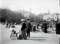 Vuxna och barn i Kungsträdgården. En kvinna med barnvagn. Några barn leker i en sandhög. Hamngatan 12 i fonden.