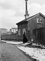 En kvinna står vid Holländarvägen 3. I fonden ses Götgatan 81 vid hörnet av Blekingegatan. Holländarvägen motsvarar ungefär nuv. Hallandsgatan (tidigare Skånegatan) och kv. Vågskivan.