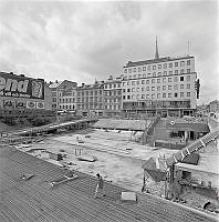 Sergels Torg under byggnad. Vy mot sydväst. T.v. brandgavlar med reklam där Kulturhuset kommer att stå. I mitten Drottninggatan 35 - 39. T.h. Klarabergsgatan.