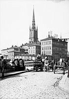 Södra sidan av Riddarholmen med Gamla Riksdagshuset och Riddarholmskyrkans torn. Vy från Munkbron med torghandel. Transport från Sankt Eriks bryggeri.