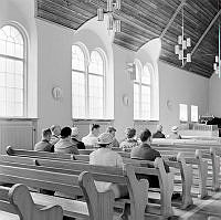 Interiör från Triangelkyrkan i Gamla Enskede. Kyrkobesökare i bänkarna.