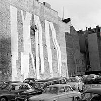 Rivningstomt med parkerade bilar i kv. Uppvaktaren. Arbetare upphissad med linor framför brandmur med plåttäckta rökgångar.