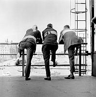 Slussen, utsikt mot Gamla Stan och Skeppsbron. Tre män står lutade mot ett räcke. Till höger en brandstege.