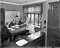 Kontorsinteriör. En rejäl Stockholmskarta, liggare, papper och penna samt ett par telefoner och ett skrivbord för fyra personer.