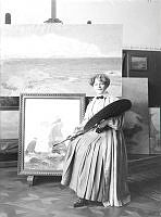 Porträtt av konstnärinnan Anna Boberg med palett framför ett staffli i ateljé.