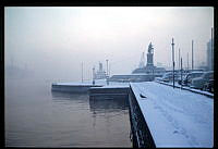 Kaj vid Skeppsbron sedd mot Gustav III:s staty. Mot söder. Till höger parkerade bilar. Vintermotiv med dis och snö.