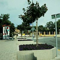 Tre trädplanteringar och en annonspelare med reklam för Kolmårdens Djurpark vid Karlaplan 11. Vy mot litet gult postkontor i kv. Fältöversten.
