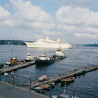 Vy från Skeppsbrons början vid Slussen mot passagerarfartyget Kungsholm för ankar på Strömmen. Bogserbåtar i förgrunden.