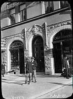 Två män står vid Odengatan 67. Till vänster en bossättningsaffär, Flodins, till höger en cigarrhandel.