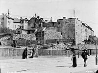 En kvinna och en man passerar ett stort vedlager vid Birger Jarlsgatan 32-30, kv. Häckelfjäll Större. Innan 1885 hette gatan Stora Träskgatan. Nuvarande Birger Jarlsgatan 34-32, kv. Sparbössan.