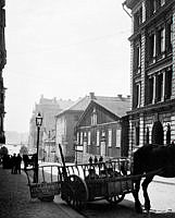 En hästkärra med mjölkkrukor står vid Grevgatan 5 (nu 15). Skylten på kärran hänvisar till S:t Paulsgatan 22. Efter trähuset t.h. ligger hörnet av Riddargatan. I fonden söderut skymtar Nybroviken.