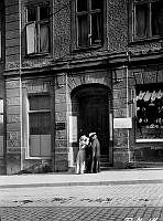 Två kvinnor står utanför porten till Hornsgatan 1. I skyltfönstret hänger löskragar och skjortbröst. Ovanför porten sitter en skvallerspegel i fönstret.