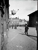 Slöjdgatan norrut vid Hötorget. En pojke kliver upp på trottoaren utanför Slöjdgatan 9. John Walls järnaffär skyltar med en vattenkanna. En kryddmakares borste och en sockertopp hänger vid nummer 11.