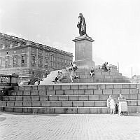 Skeppsbron, Gustav III:s staty. Sittande människor på trappstegen. I bakgrunden Stockholms slott.