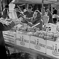 Fruktförsäljning på Hötorget. En torgmadam med sjalett står vid en rad äppellådor med texten 
