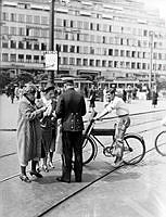 Fyra trafikdagar i Stockholm där Trafikfrämjandet la tonvikten på cyklismen som utgör en växande trafikfara. Artiga Poliskonstaplar delar ut broschyrer om cykelvett vid Norrmalmstorg mot Citypalatset.