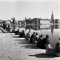 Stadshusträdgården. Stockholmare har i vårvärmen sökt sig till stadens vatten och slagit sig ner vid Stadshuset. Vy mot Riddarholmen. Skulpturen till vänster är Carl Eldhs 