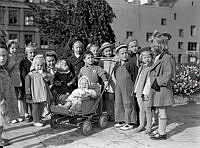 Grupporträtt av en samling barn vars mammor vid tillfället ägnar sig åt husmodersgymnastik. Här passas de av barnträdgårdslärarinnan Margot Fränckels.