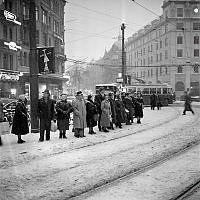 Stureplan och Birger Jarlsgatan söderut. Stureplan 2 och 3. Folk väntar på buss/spårvagn i snöyran. På två stolpar sitter affischer med ett snapsglas och ett ljus, 