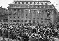 Gustav Adolfs Torg, mot kvarteret Vinstocken - Skandinaviska Banken. Folksamling ser på Norges kung Haakon och Gustaf VI Adolf i kortege genom Stockholm under kung Haakons officiella besök.