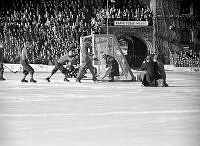 Stadion. SM-finalen i Bandy mellan Nässjö och Edsbyn. Högerytter Uno Wennerholm (nr 7) skjuter direkt på tennisslag och bollen tar just ovanför hatten på den hukande måldomaren.